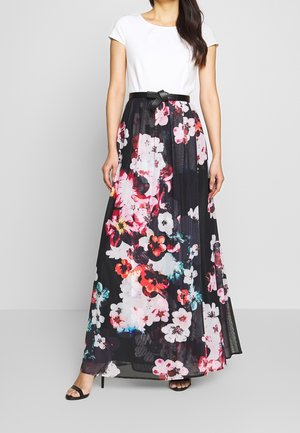 Kobieta ubrana w biały top z krótkim rękawem oraz czarną długą spódnicę w kwiaty z czarnym paskiem, do tego czarne sandały na obcasie z odkrytymi palcami.