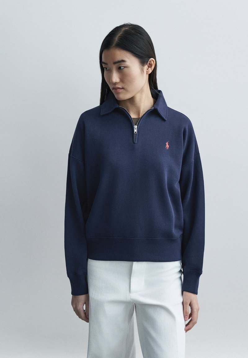 Tumesinine lukuga dressipluus kraega ja väikese punase logoga. Pehme tekstuur, lai lõige ja soonikkoes varrukasäärised. Kandmiseks koos valgete pükstega.