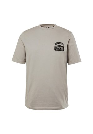 Jasno beżowa bawełniana koszulka z luźnym krojem. Posiada czarną naszywkę na piersi z tekstem i szczegółami graficznymi. Okrągły dekolt i krótkie rękawy.