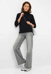 Zwarte top met lange mouwen en een col, gecombineerd met lichtgrijze jeans met wijde pijpen en zilveren sneakers. Model draagt een zilveren schoudertas.