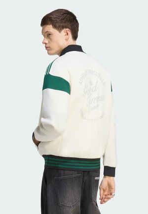 Jonge man draagt een wit-groene Adidas-jas met tekst op de achterkant, staat met handen in de zakken, kijkt iets naar links.
