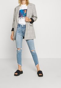 Šedý kostkovaný oversize blejzr přes bílý tričko s grafickým potiskem, doplněný o obnošené světlé modré džíny a černé klouzavé sandály.