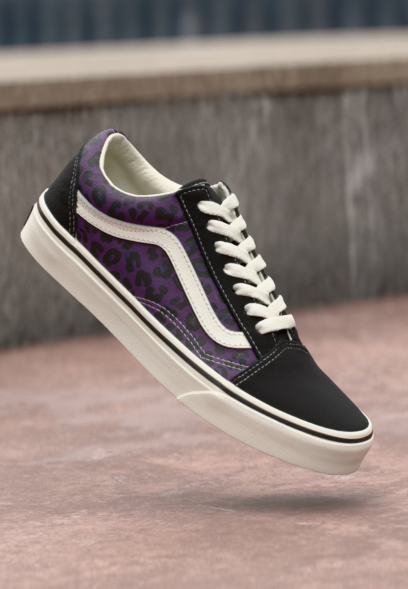 Basket imprimée léopard noire et violette avec lacets et semelle blancs, flottant au-dessus d'une surface rosée avec un arrière-plan gris flou.
