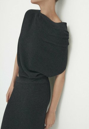Femme portant une robe en maille gris foncé ajustée avec un large décolleté drapé, se tenant devant un fond blanc uni.