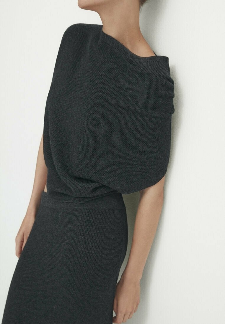 Femme portant une robe en maille gris foncé ajustée avec un large décolleté drapé, se tenant devant un fond blanc uni.