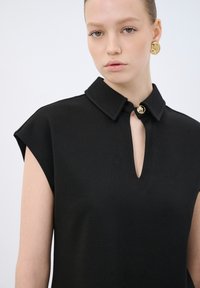 Vestito nero a maglia con colletto corto, maniche a cappuccio e un'apertura a forma di chiave sul davanti con un bottone dorato decorativo. Tessuto liscio.