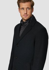 Zwarte gekleurde jas met een notch lapel, voorzien van grote zwarte knopen en een verborgen zwarte ritssluiting onder de jas.