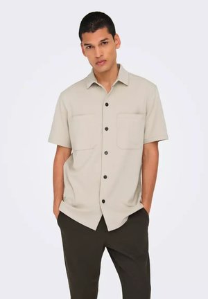 Camisa beige de manga corta con cuello tipo button-down, dos bolsillos en el pecho y botones negros; llevada con pantalones oscuros sobre un fondo claro.