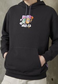 Sweat à capuche noir avec poche avant et personnages de dessins animés colorés jouant au basketball, baseball et tennis imprimés au-dessus de la poche.