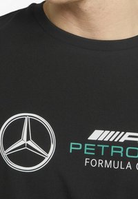 Mustapaita pyöreällä kaula-aukolla, jossa on valkoinen Mercedes-logot ja tealsininen "PETRONAS F1" -teksti, sileä kankaan rakenne.