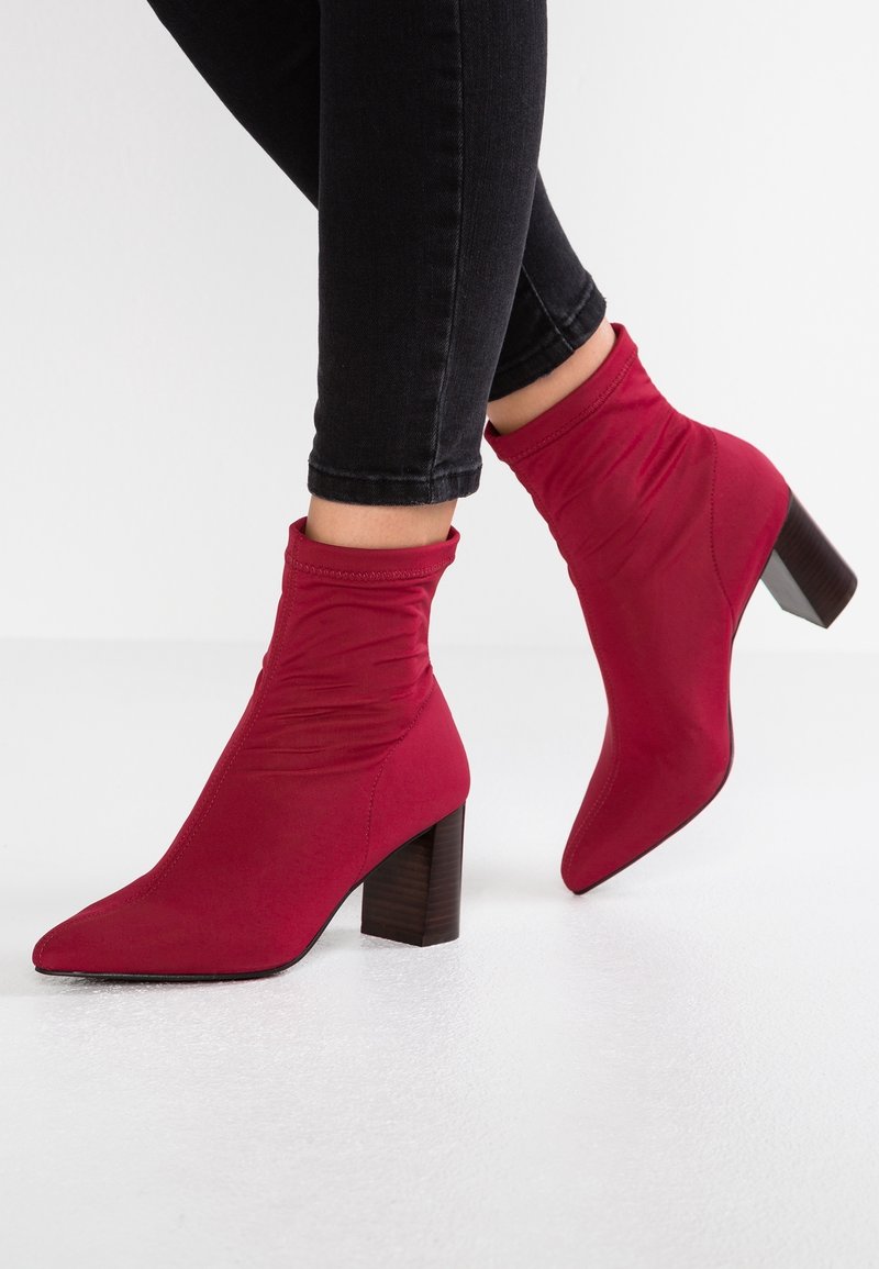 Personne portant un jean skinny noir et des bottines rouges à talons blocs sur une surface blanche.