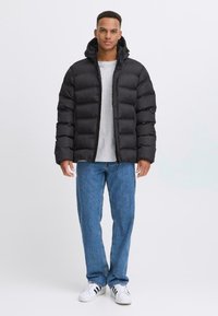 Sort puffjakke med hætte, der har en quiltet design. Bæres over en grå skjorte og kombineres med blå jeans og hvide sneakers.