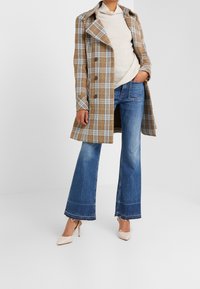 Persoon in een beige geruite jas, crèmekleurige ribbeltrui, blauwe cropped flare jeans en beige puntige high heels staat op een witte achtergrond.