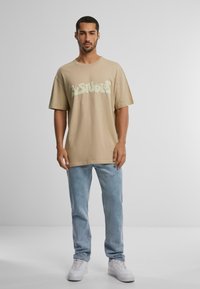 Beige oversized t-skjorte med lysegrønn grafisk tekst, kombinert med lyseblå jeans og hvite sneakers. Minimalistisk design mot en nøytral bakgrunn.
