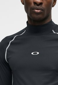 Κοντινό πλάνο ενός ατόμου που φοράει μαύρο αθλητικό πουκάμισο με ψηλό γιακά, λευκές ραφές και λογότυπο Oakley στο στήθος.