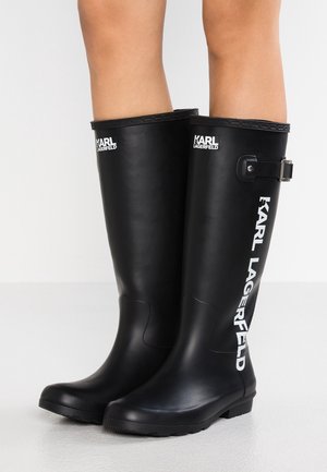 Lange zwarte rubberen laarzen met "KARL LAGERFELD" branding op de voorkant en zijkant, gedragen op blote benen tegen een effen achtergrond.