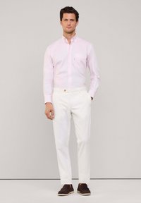 Camicia button-up rosa chiaro con tasca sul petto, abbinata a pantaloni bianchi e scarpe slip-on marroni. Texture liscia e linee pulite.