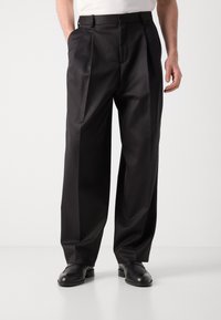 Schwarze plissierte Hose aus glattem Stoff, mit hoher Taille, seitlichen Taschen und tailliertem Schnitt bis zu den Schuhen.