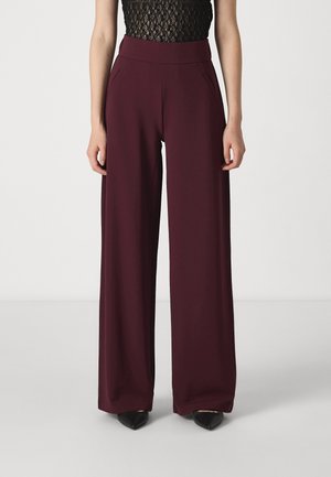 Femme portant un pantalon bordeaux taille haute à jambes larges avec poches latérales, associé à un haut en dentelle noire et des chaussures noires à bouts pointus.