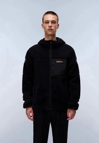 Jovem vestindo uma jaqueta de fleece preta com zíper e capuz, com um bolso de patch no peito, posando contra um fundo claro e liso.
