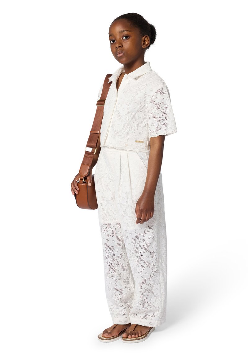 Witte kanten jumpsuit met korte mouwen, bloemenpatroon en tailleband. Gecombineerd met een tan crossbody tas en bruine slippers.