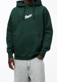 Personne portant un sweat à capuche vert foncé avec le logo blanc "SWT." et un jean gris clair, une main dans la poche, sur un fond blanc uni.