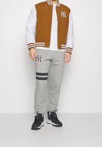 Brun varsityjacka med vita ärmar och ränder, grå träningbyxor med svarta ränder, broderad logo och svarta joggingskor.