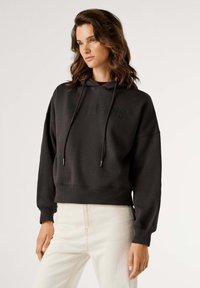 Felpa corta nera con coulisse e testo frontale visibile. Realizzata in tessuto morbido con polsini e orlo a coste, abbinata a pantaloni chiari.