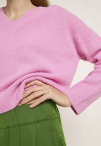 Rosa gestrickter Pullover mit V-Ausschnitt und gerippten Bündchen, getragen über einem grünen Faltenrock. Der Stoff wirkt weich und strukturiert.