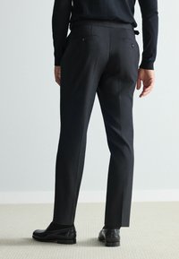 Schwarze Anzughosen mit maßgeschneidertem Schnitt, ausgestattet mit glatter Textur, Gesäßtaschen und dezenten Details, kombiniert mit glänzenden schwarzen Schuhen.