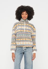 Fliisist pullover kõrge kraega, mitmevärviliste geomeetriliste mustritega, elastse ääre ja nööriga. Pehme tekstuur; põhivärvid on oranž, sinine ja roheline.