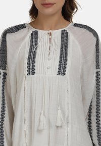 Biała bluzka z czarnymi haftowanymi wzorami, z przodem zapinanym na guziki, sznurkiem do ściągania oraz plisowanym wykończeniem na korpusie i rękawach.