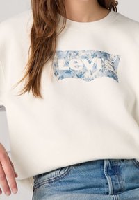 Sudadera blanca con un logo floral azul de Levi's en el frontal. Textura suave, diseño de cuello redondo, combinada con jeans de mezclilla azul claro.