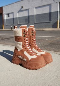 Paar hoher, braun-weißer Winterstiefel mit Fleecefutter und Schnürung, mit dicken Sohlen, stehend auf einem Bürgersteig vor einem urbanen Straßenhintergrund.