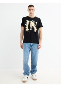 Camiseta de algodón negra con un diseño abstracto crema en la parte delantera, combinada con jeans de mezclilla azul claro y zapatillas blancas con detalles en marrón.