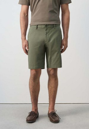 Uomo che indossa pantaloncini verde oliva fino al ginocchio, scarpe marroni con lacci e una camicia marrone a maniche corte, in piedi su un pavimento grigio contro una parete semplice.
