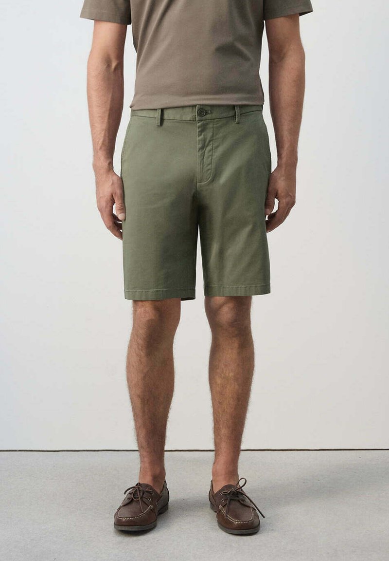 Homme portant un short vert olive au genou, des chaussures marron à lacets et une chemise marron à manches courtes, debout sur un sol gris devant un mur uni.