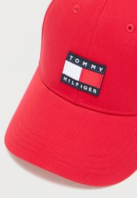 Κόκκινο καπέλο μπέιζμπολ από βαμβάκι με καμπυλωτό γείσο. Έχει μια μαύρη ετικέτα με την επιγραφή "TOMMY HILFIGER" και ένα ορθογώνιο λογότυπο σε λευκό, κόκκινο και μπλε.