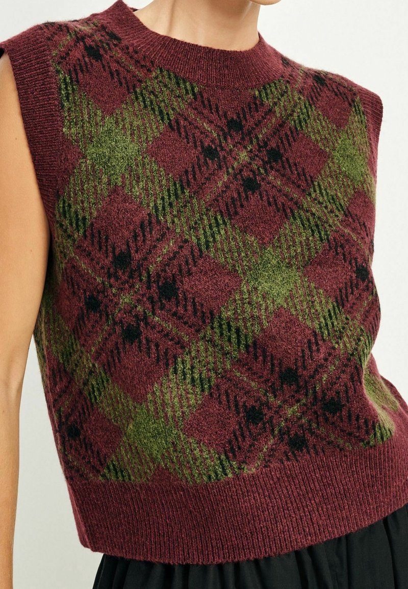 Gilet en maille sans manches avec motif losange vert et noir sur fond bordeaux, porté sur une jupe noire.