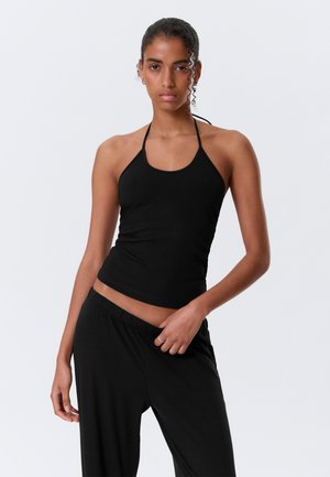 Femme aux cheveux bouclés portant un haut dos nu noir et un pantalon noir, posant avec une main dans la poche sur un fond clair uni.