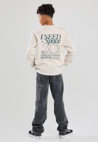 Cream sweatshirt with large green graphic text on back reading "I NEED Space" and geometric designs. Paired with gray jeans and black sneakers.