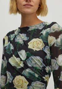 Langärmliges Kleid aus schwarzem Stoff, geschmückt mit großen weißen und cremfarbenen Rosen. Das Material ist transparent und hat ein drapiertes Design.