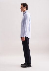 Camicia a maniche lunghe azzurro chiaro con colletto a bottoni, taschino sul lato sinistro del petto, abbinata a pantaloni scuri e scarpe nere.