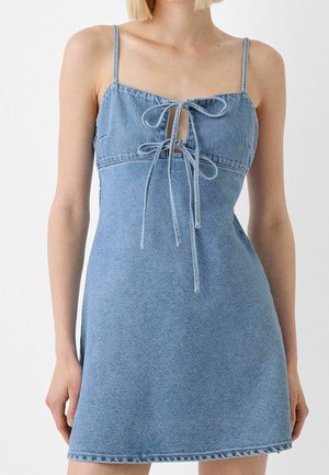 Mujer con vestido mini azul de mezclilla sin mangas, con dos lazos frontales y tirantes de espagueti, sobre un fondo liso.