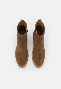 Botas de tobillo de ante marrón con una textura suave, punta redonda y un tacón bajo. Presentan una cremallera lateral y una pestaña en la parte posterior para facilitar su puesta.