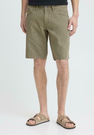 Mężczyzna w khakich, casualowych szortach i beżowych sandałach z odkrytymi palcami, stojący na jasnym, jednolitym tle.