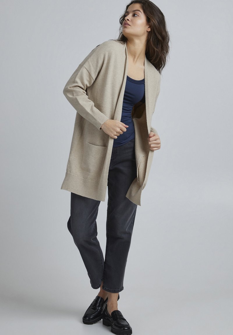Beige Strickjacke mit offenem Vorderteil, langen Ärmeln und zwei vorderen Taschen, kombiniert mit einem marineblauen Oberteil und dunkelgrauen Jeans, vor einem neutralen Hintergrund.