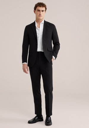 Noor mees, kes kannab musta istuvat ülikonda koos valge triiksärgiga ja mustade kingadega, seisab ühe käega taskus tavalise tausta ees.