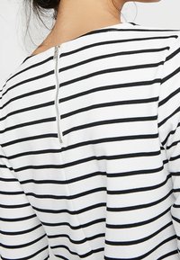 Raidallinen paita, jossa mustavalkoiset vaakasuorat raidat, pyöreä pääntie ja näkyvä hopeinen vetoketju takana. Pehmeä kangaspinta.