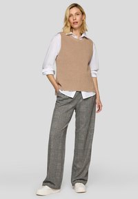 Beige gerippter, ärmelloser Strickpullover, der über einem weißen Hemd mit Knöpfen getragen wird, kombiniert mit grauen, karierter, weit geschnittenen Hosen und weißen Turnschuhen.